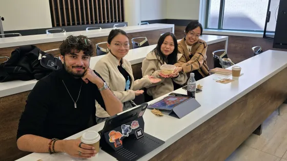 Four students sit at a table, laptops and food items in front of them. 