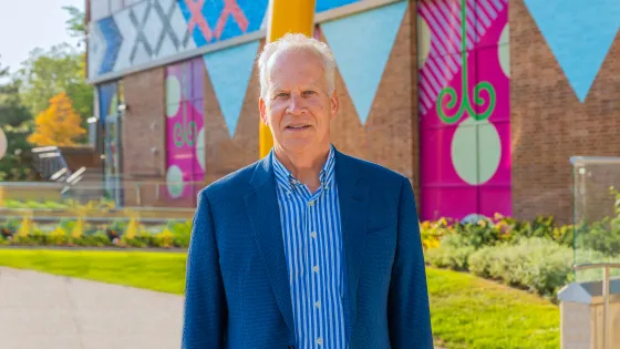 Photograph of David Binet wearing a blue blazer, standing in Butterfield Park on a sunny day.