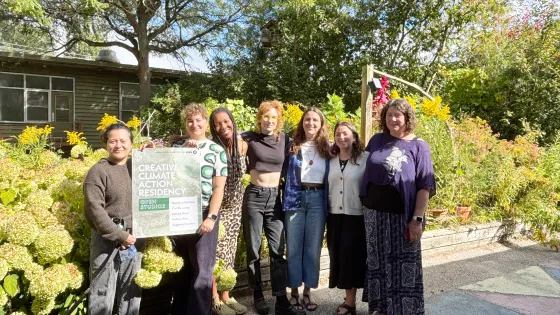 A group of seven people standing in front of a building with green bushes in the background
