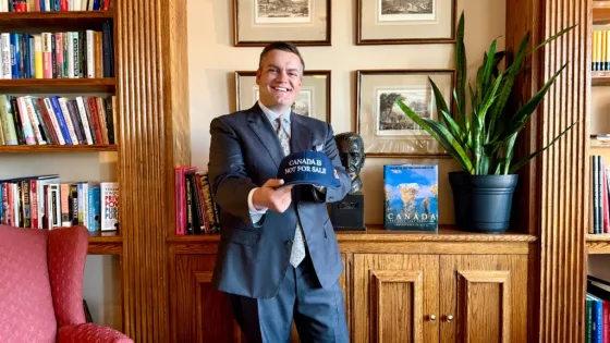 Photo of a man wearing a blue suit and tie holding up a baseball cap that says Canada is not for sale, standing in front of a cabinet with bookshelf to the left, and photos hanging on the wall behind him.