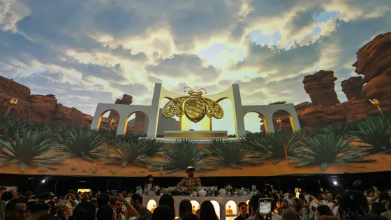 An event with a DJ performing on stage with a screen in the background displaying a desert scene, architectural structures and a golden bee in the center.