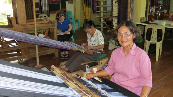 Three weavers working on a loom, woman in foreground smiling at camera wearing pink shirt