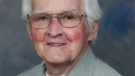 Photo of a man with glasses and white hair wearing a grey shirt with a white t-shirt underneath