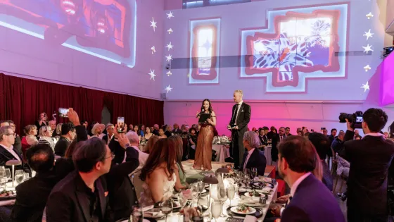 Gala Co-Chairs Amy Burstyn-Fritz and Jeff Hull stand on the centre stage addressing guests during the 2026 OCAD U Gala. Attendees seated at banquet tables surround the stage while large-scale projected visuals from Solarium by OCAD U alum Cell Driver illuminate the walls of the Great Hall.