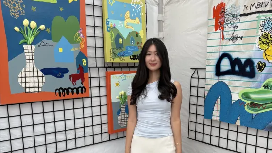 Woman with long black hair wearing pale blue blouse and white skirt standing among her paintings