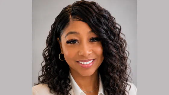 Woman with long dark curly hair, wearing white shirt and beige blazer
