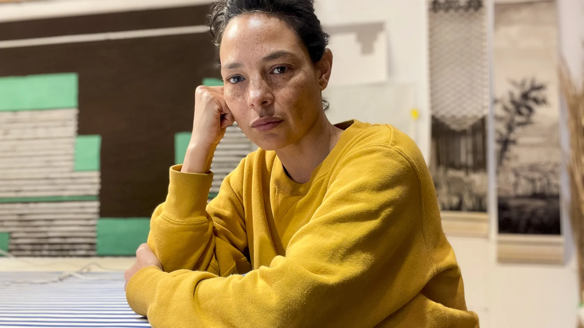 Photo of Christine Howard Sandoval sitting at a table, wearing a yellow sweater and with her arms on the tabletop.