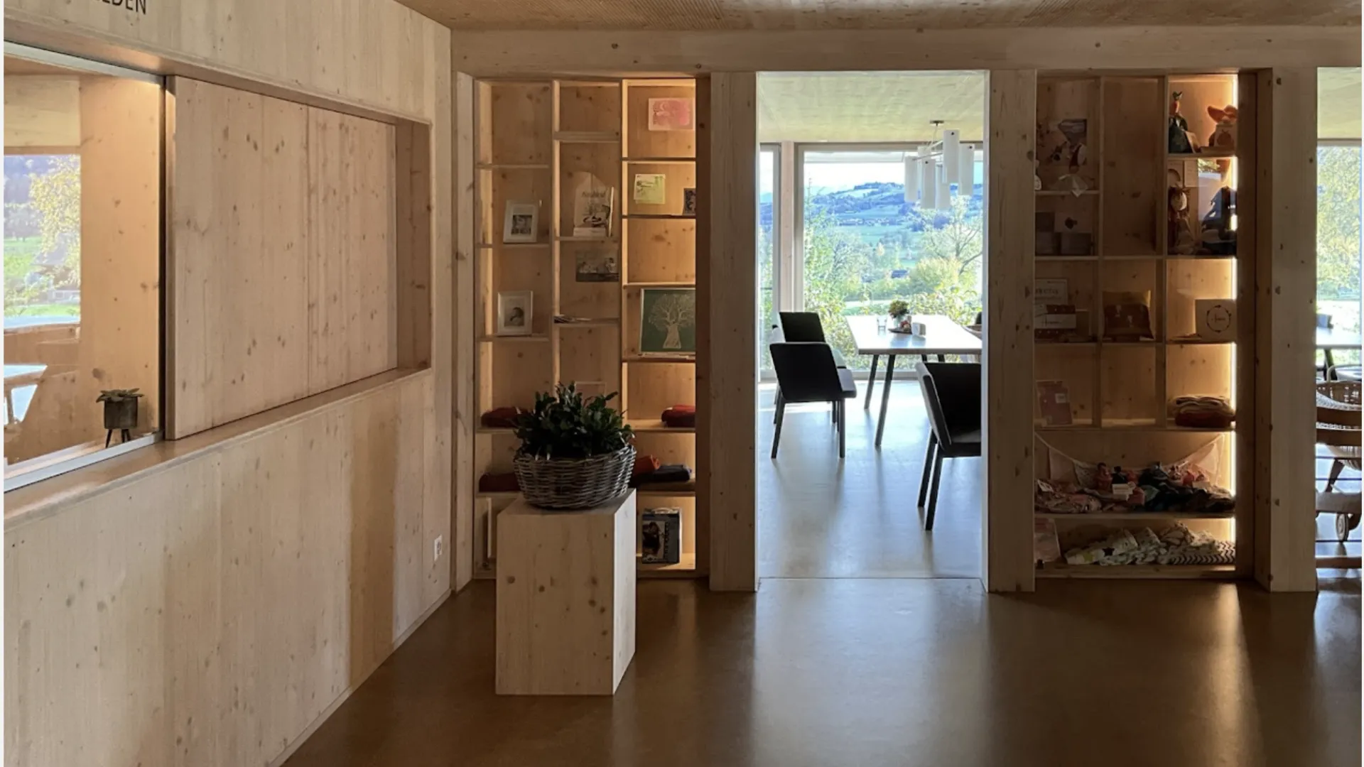 Photo of a room with wooden interiors. There are shelfs with decorative things on them. There is a table and a couple of chairs in the next room, which also has a wall to wall window.