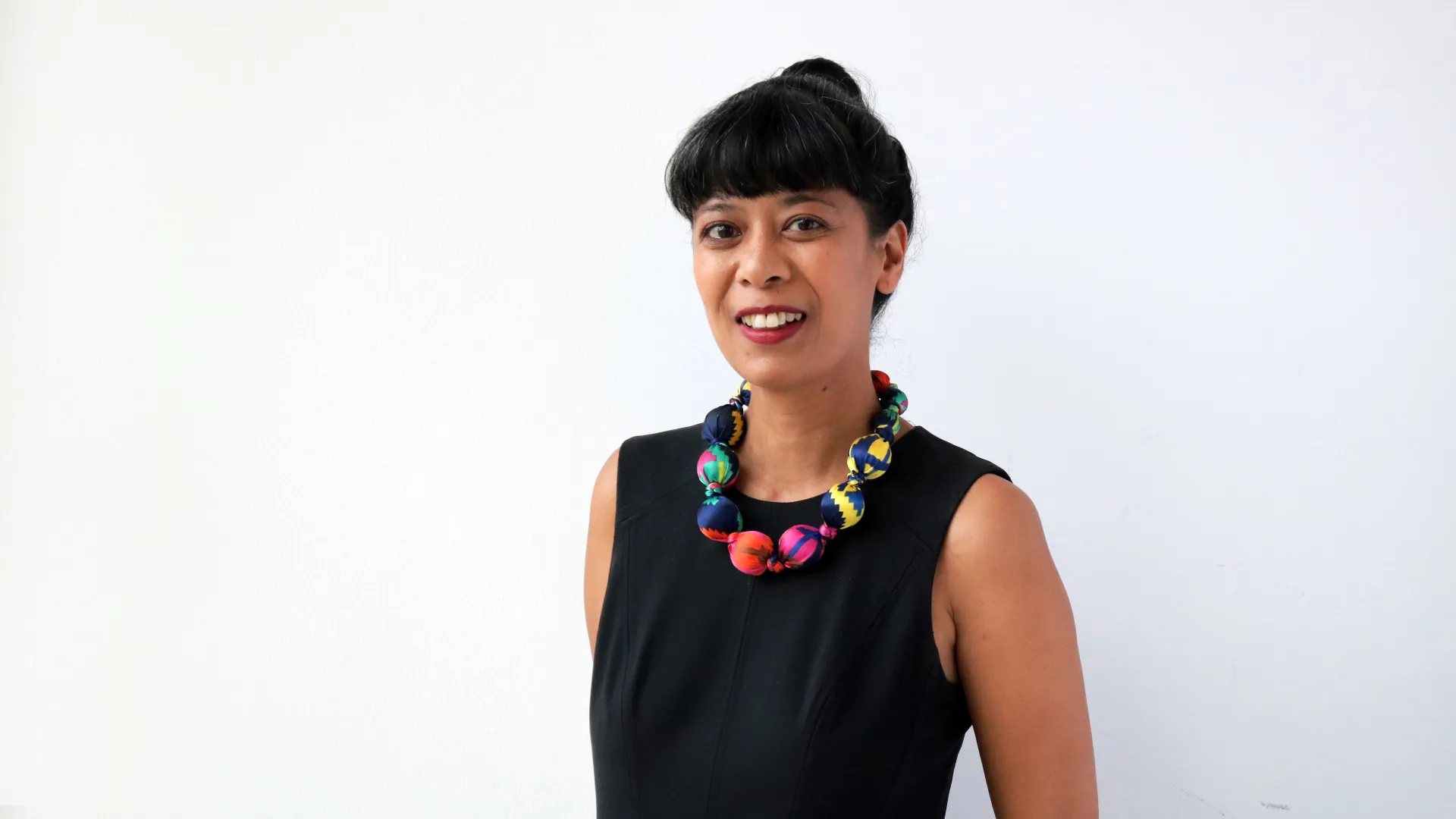 A woman with black hair in a bun wearing sleeves black shirt and colourful necklace