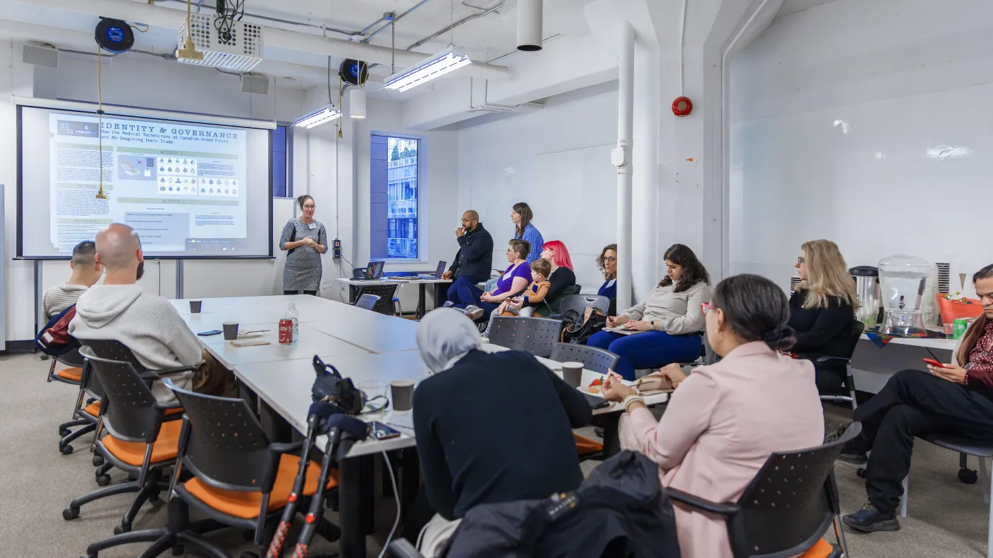 White room with a table in the centre, while people look towards a woman.