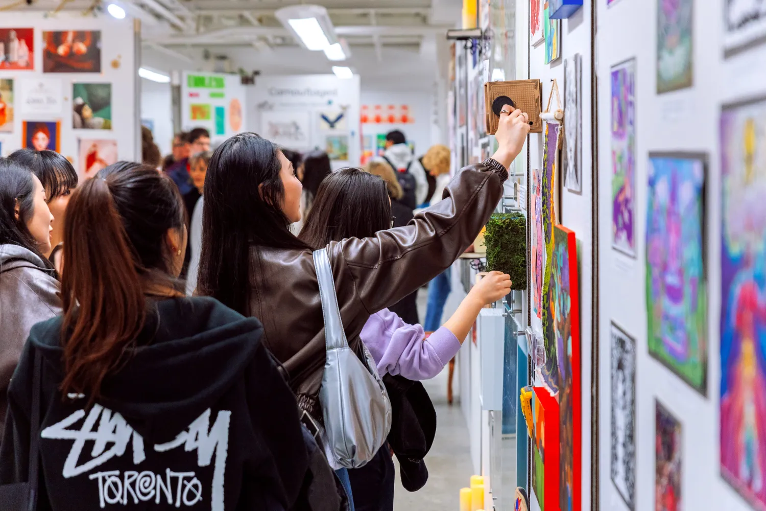 People at an art show reaching up to a white wall filled with small artworks