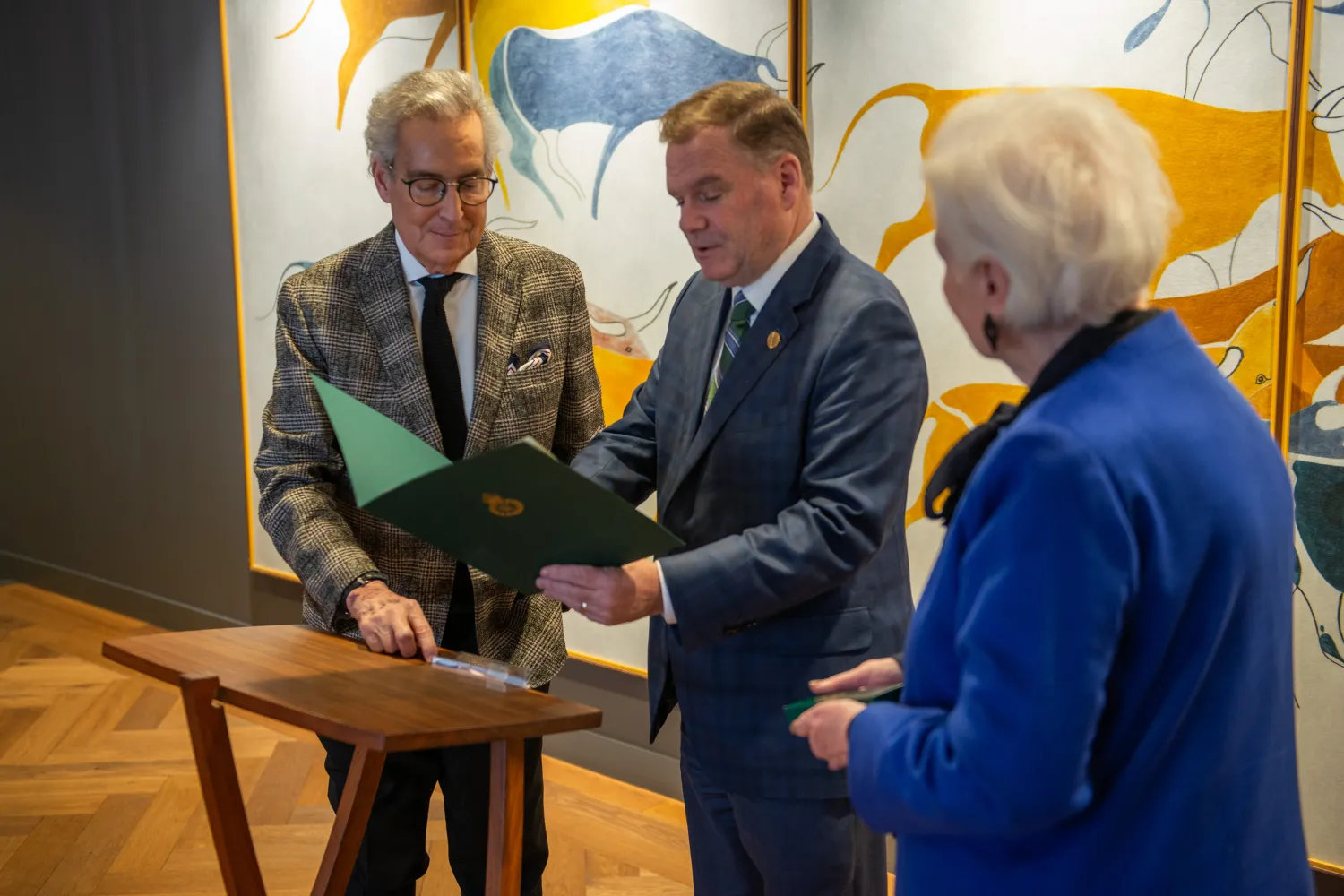 Three people in photo, at left a man with white hair and glasses wearing a grey patterned suit, man in middle holding a certificate and at far right woman with white hair wearing blue suit