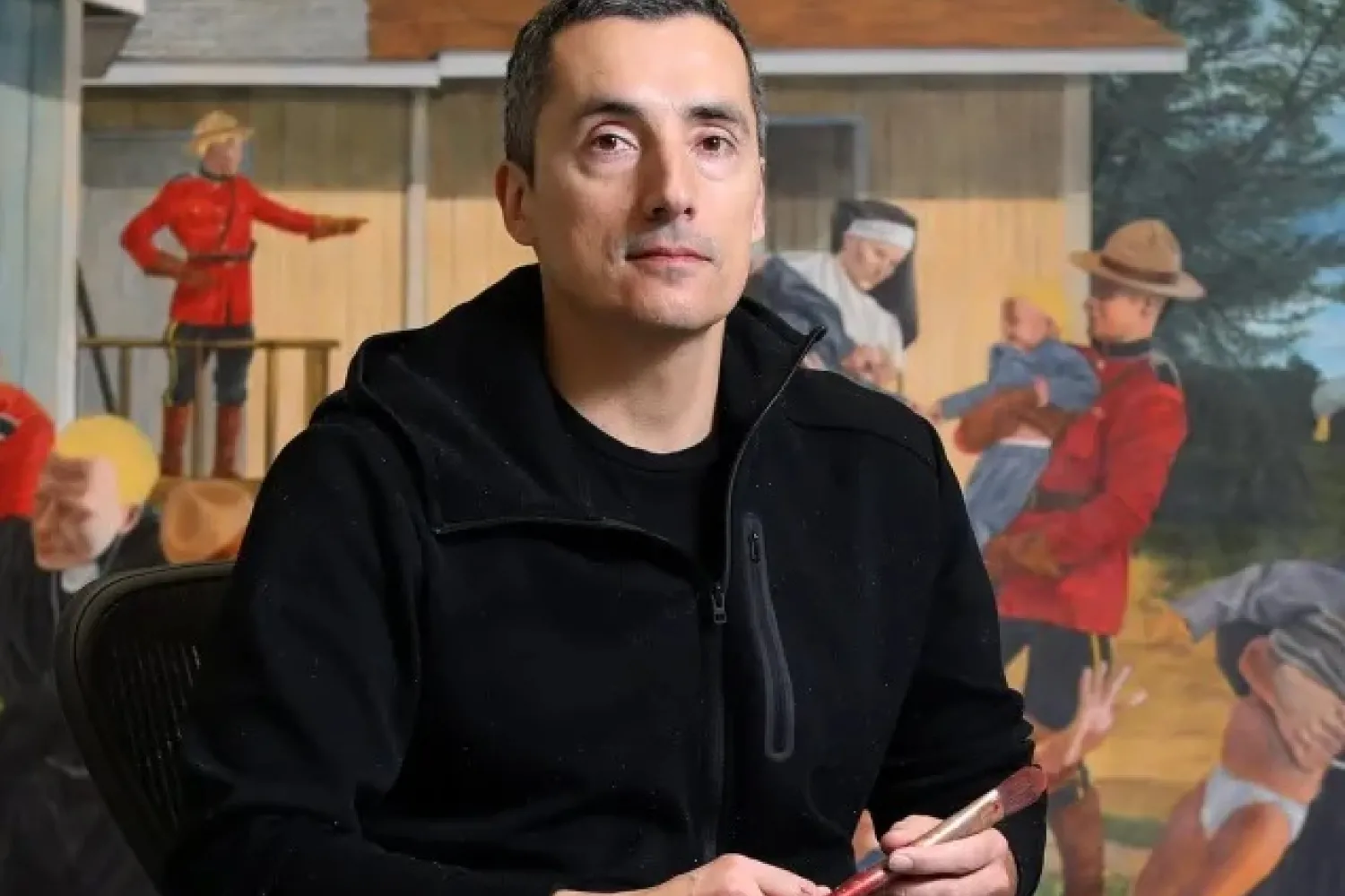 Colour photo of Kent Monkman holding a paintbrush, seated in front of his art work. 