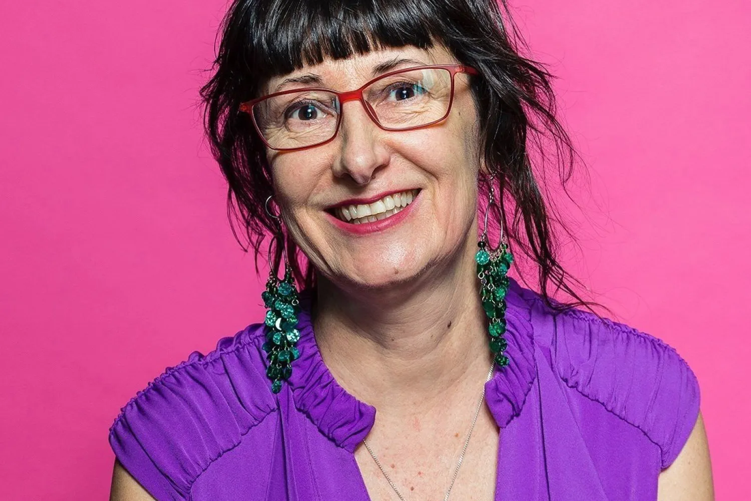 A woman with brown hair, brown eyes wearing red eye glasses, green earrings, purple shirt