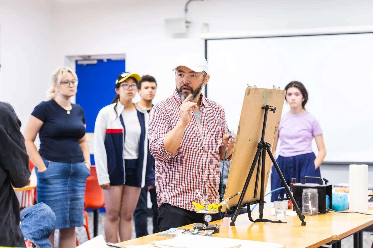 Students gather around an instructor giving an art demonstration in a studio space. Attention is focussed around an easel placed on top of a table. 
