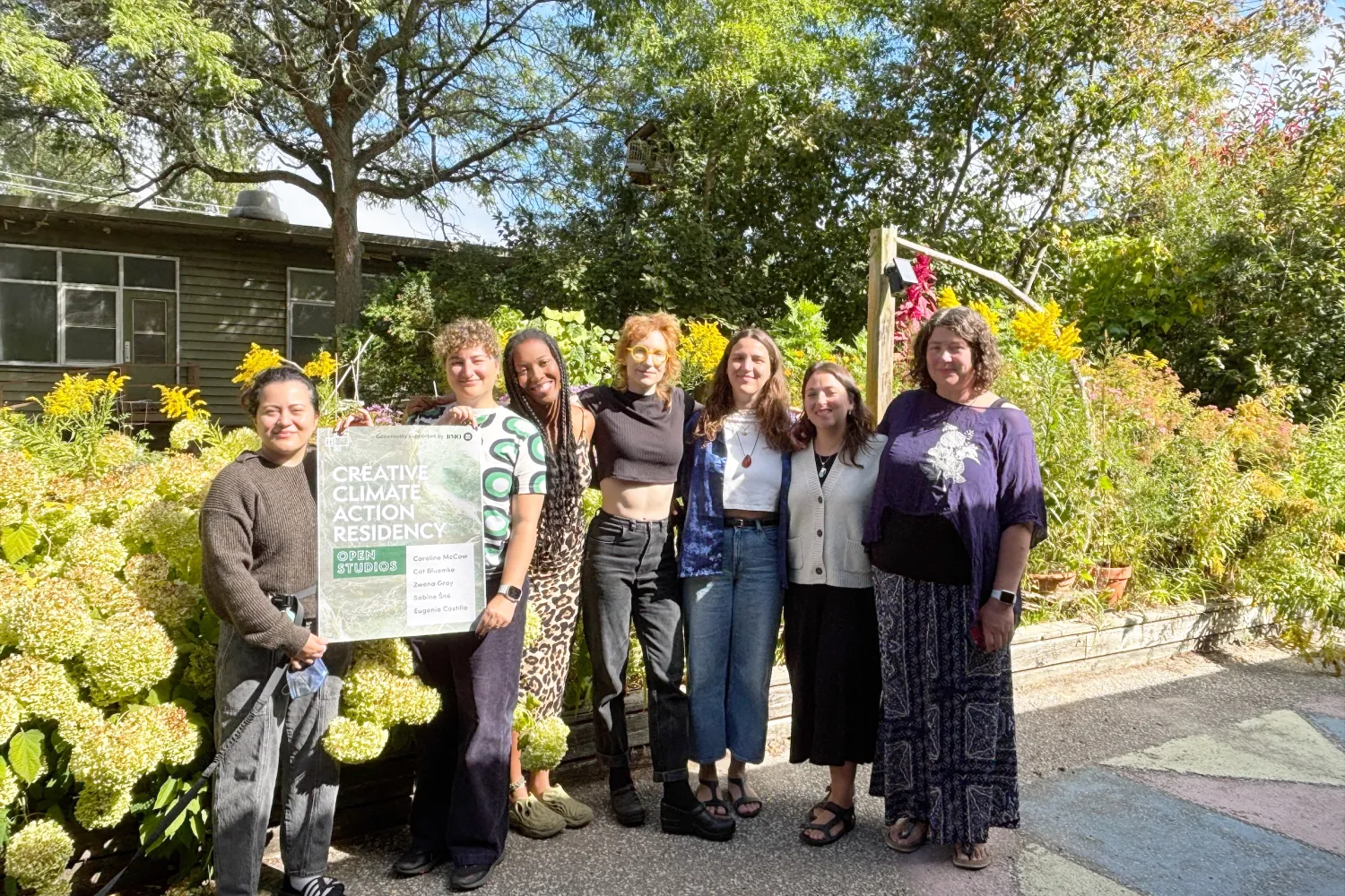 A group of seven people standing in front of a building with green bushes in the background