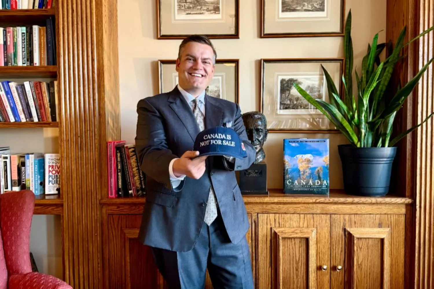 Photo of a man wearing a blue suit and tie holding up a baseball cap that says Canada is not for sale, standing in front of a cabinet with bookshelf to the left, and photos hanging on the wall behind him.