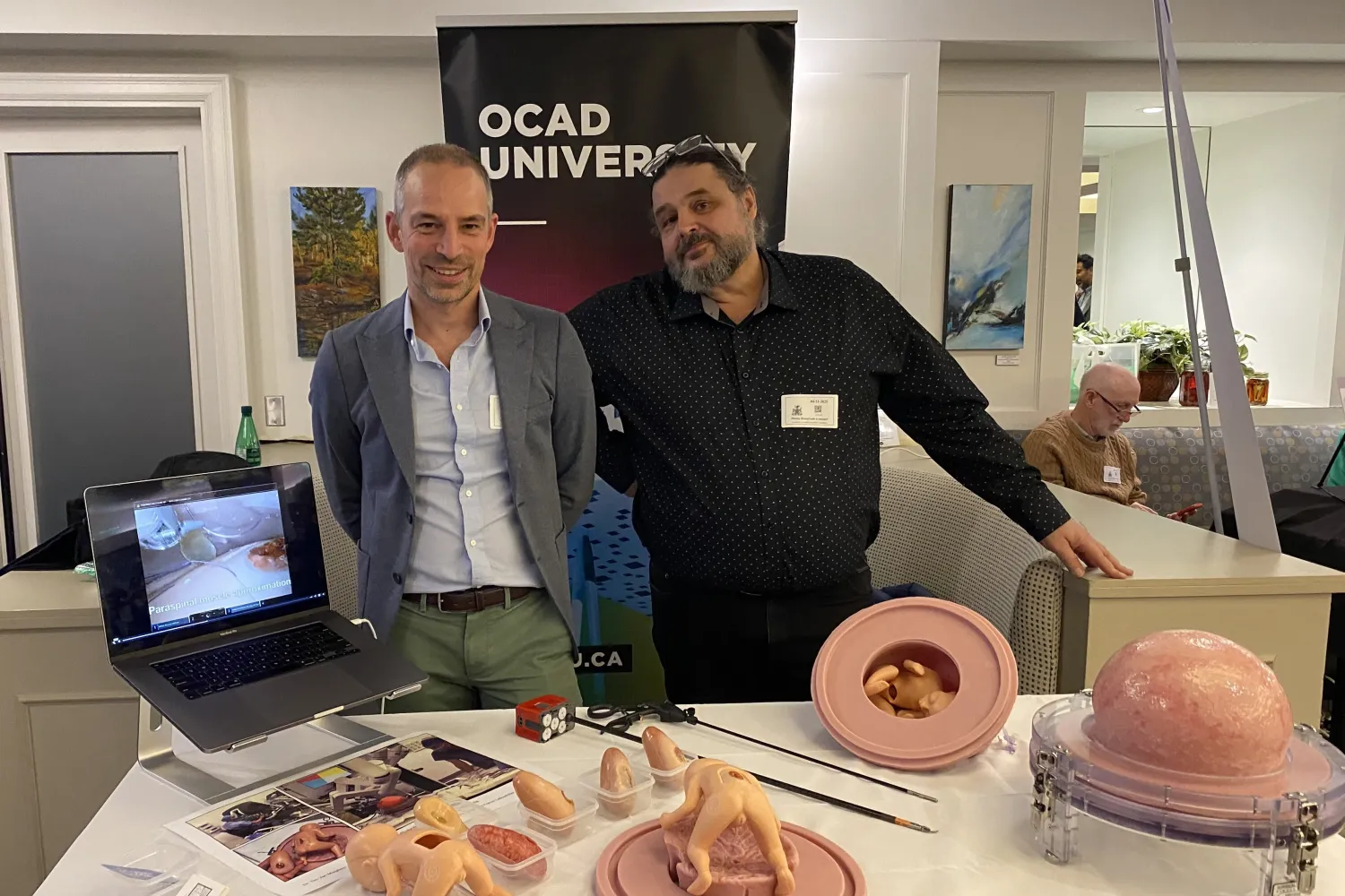 Dr. Tim Van Mieghem and Professor Francis LeBouthillier, standing in front of a table displaying medical surgical models, with an OCAD U banner in the background. 