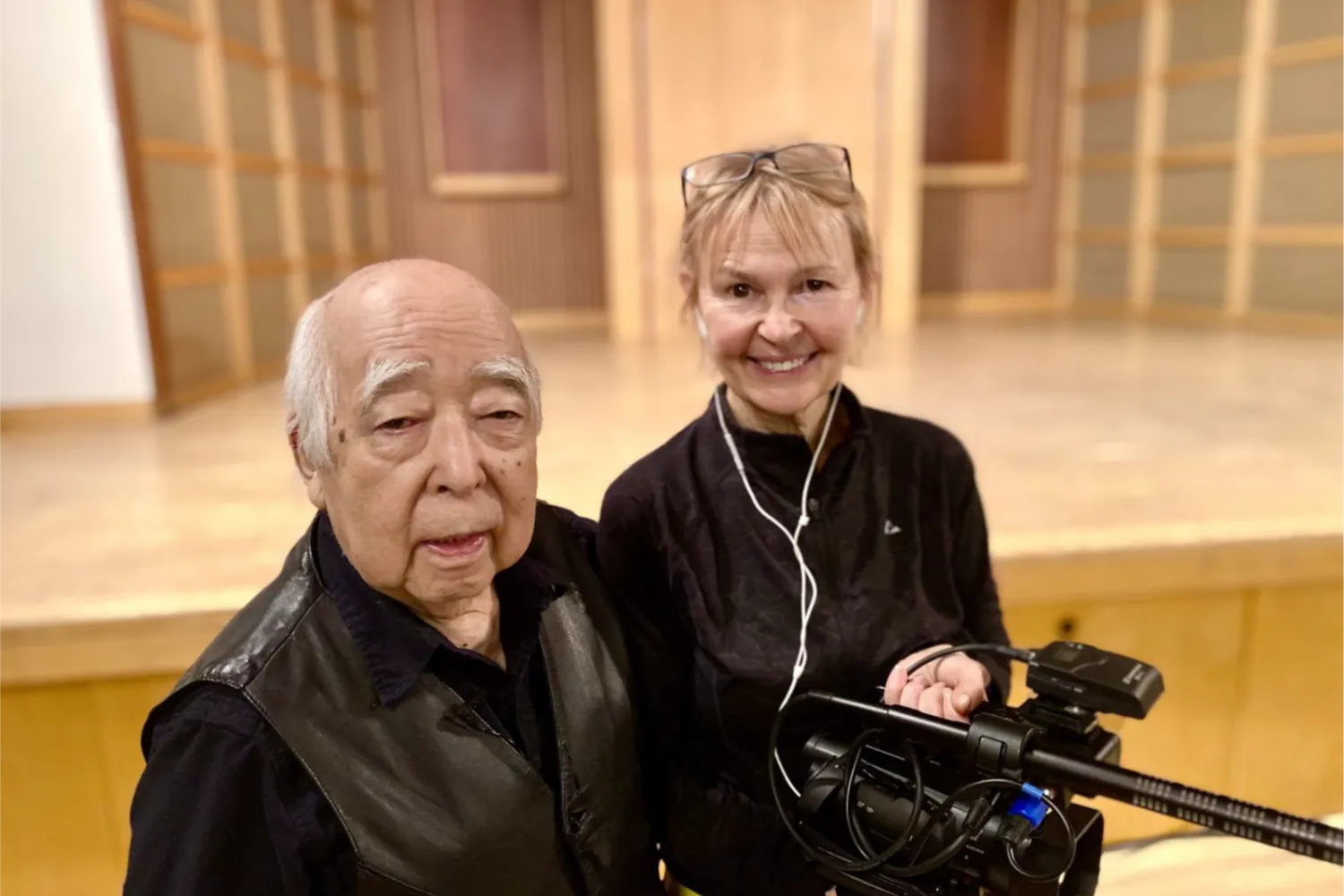 Japanese-Canadian man at left with a woman with blond hair and glasses on her head, holding a camera.