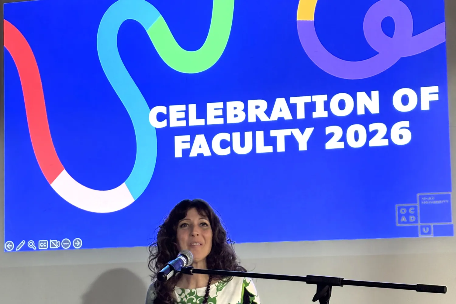 Provost Dr. Sandra Gabriele standing at podium with microphone, background slide of blue with colourful swirl saying, Celebration of Faculty 2026