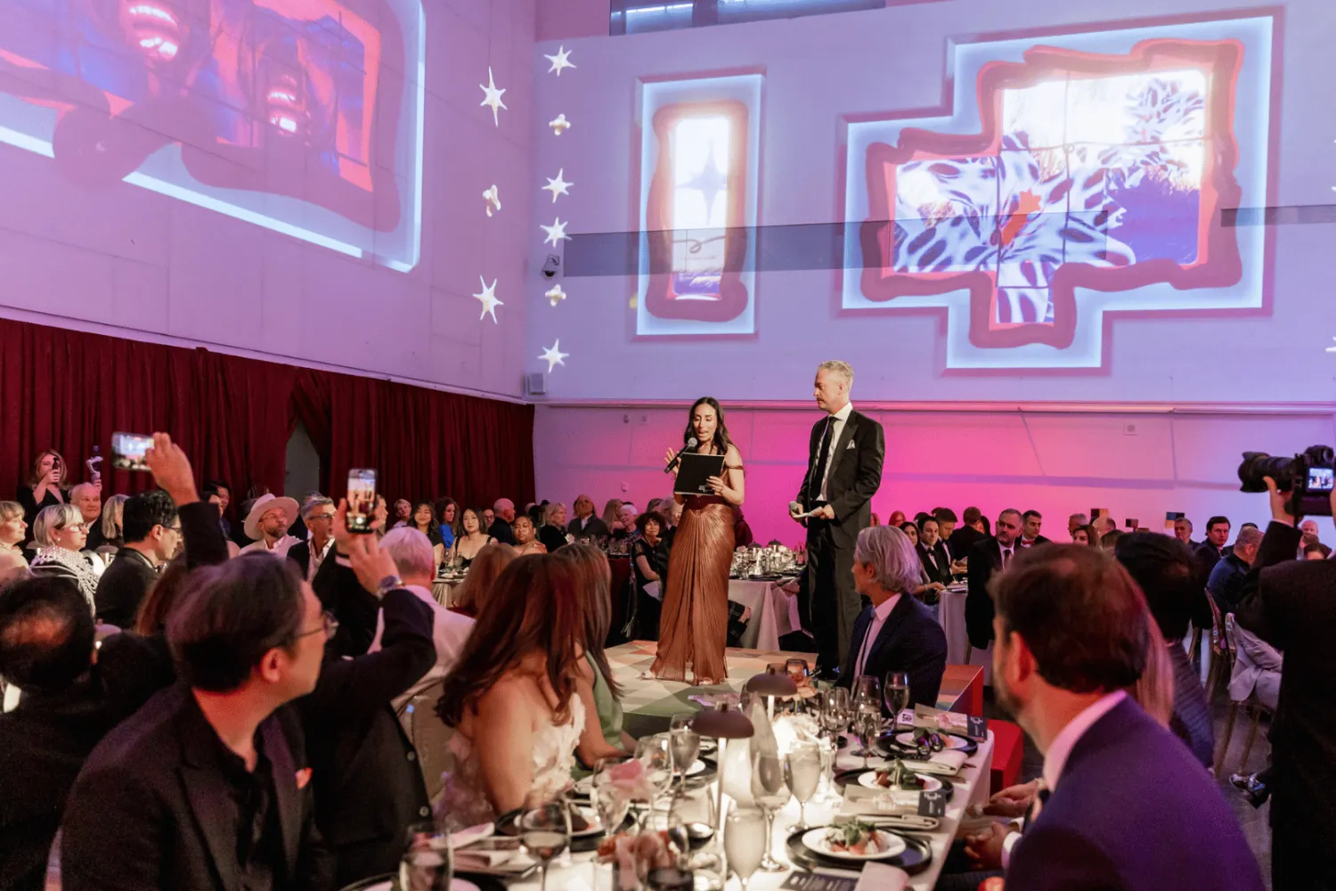 Gala Co-Chairs Amy Burstyn-Fritz and Jeff Hull stand on the centre stage addressing guests during the 2026 OCAD U Gala. Attendees seated at banquet tables surround the stage while large-scale projected visuals from Solarium by OCAD U alum Cell Driver illuminate the walls of the Great Hall.