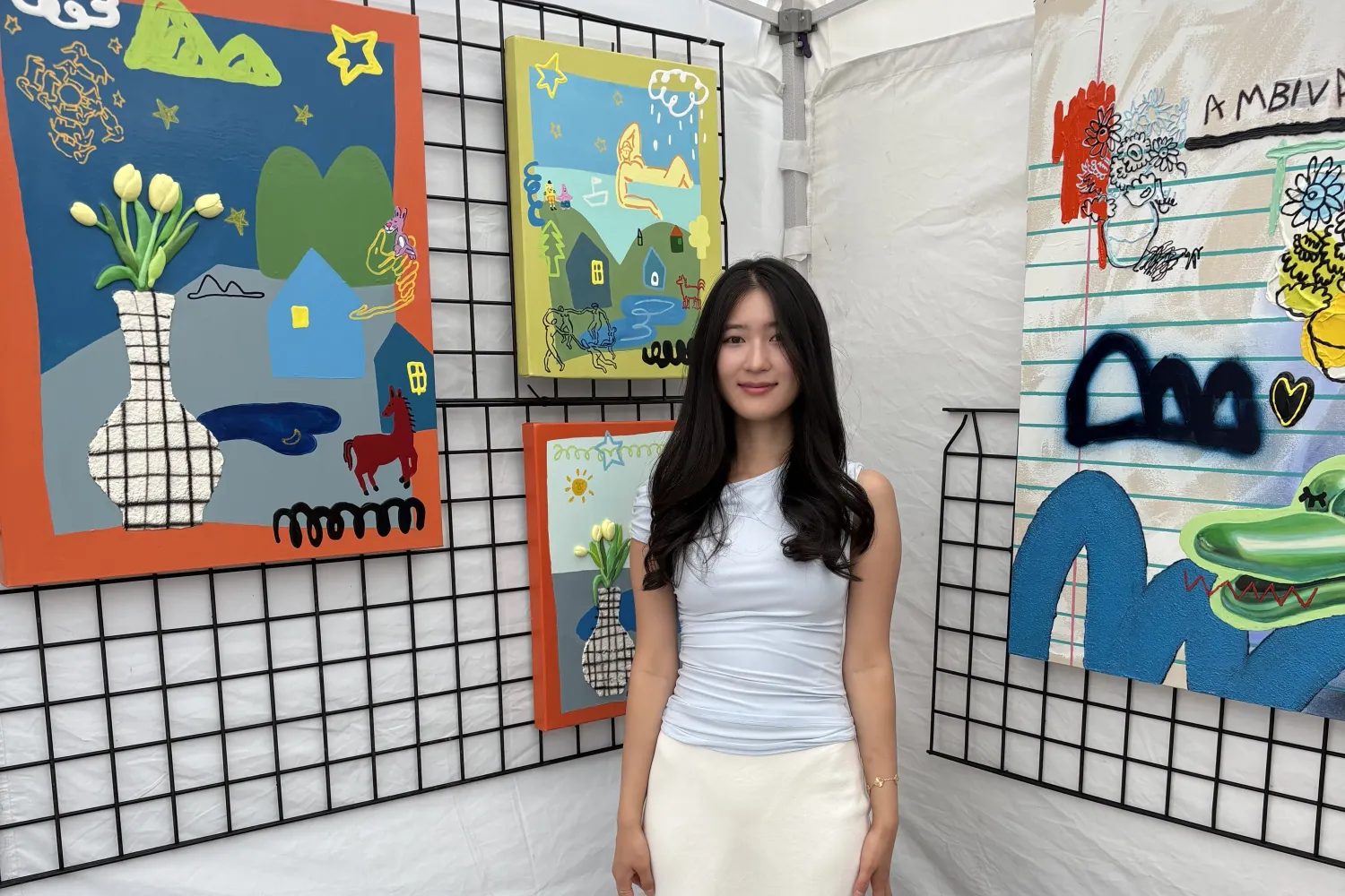 Woman with long black hair wearing pale blue blouse and white skirt standing among her paintings