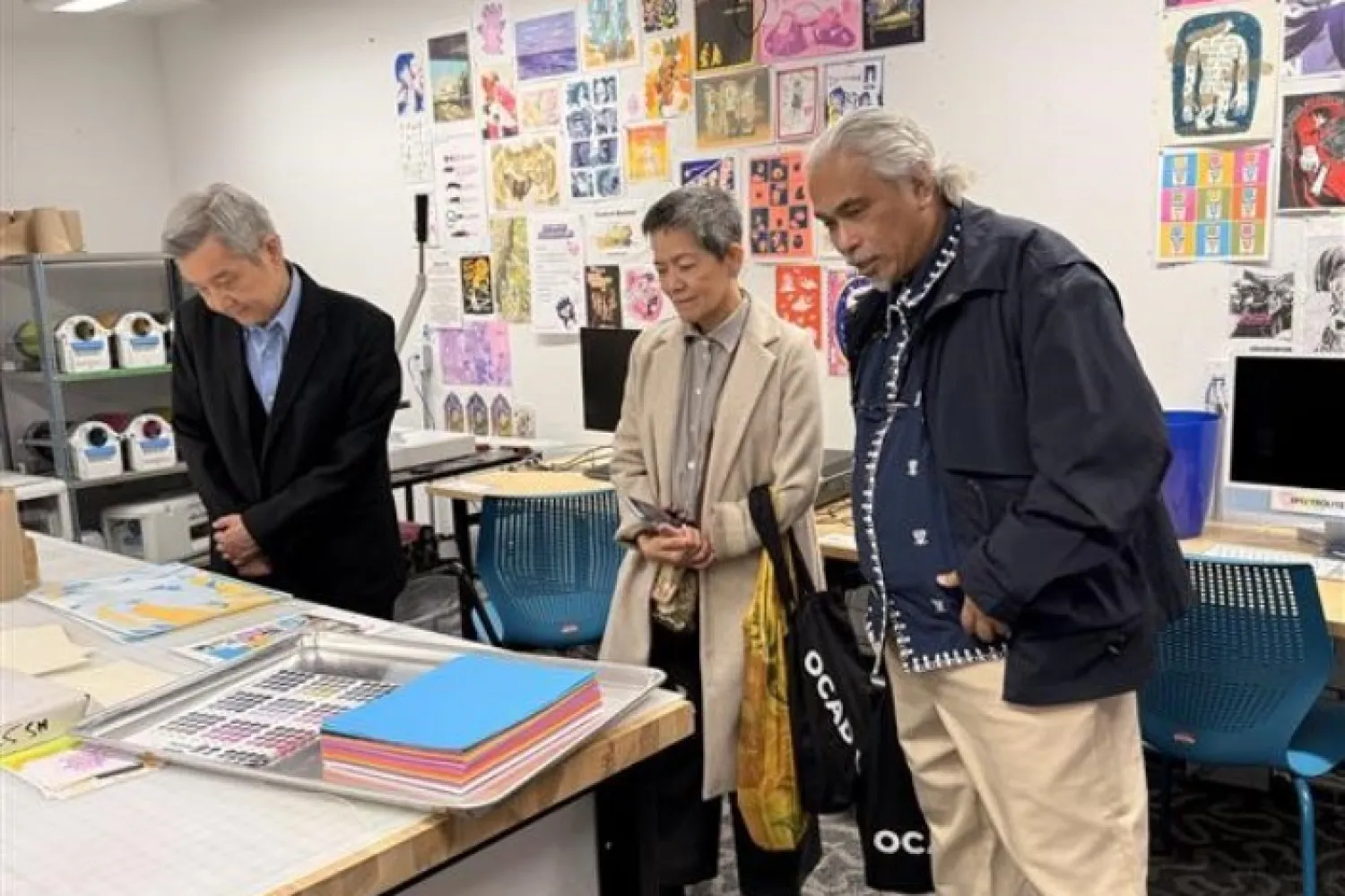 Three people looking at artwork. A man (on the left) wearing a black suit; a woman (in the middle) wearing a beige coat; and another man (on the right) wearing a black jacket and beige pants. 
