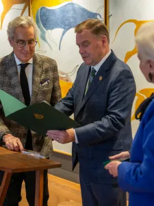 Three people in photo, at left a man with white hair and glasses wearing a grey patterned suit, man in middle holding a certificate and at far right woman with white hair wearing blue suit