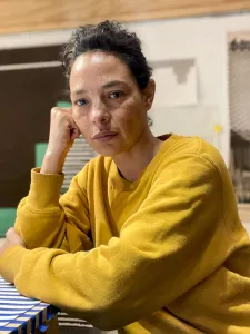 Photo of Christine Howard Sandoval sitting at a table, wearing a yellow sweater and with her arms on the tabletop.