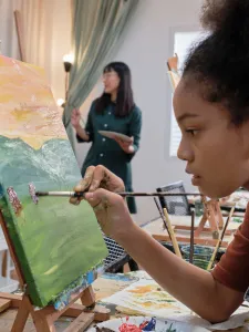 Young female teen is painting at an easel and an instructor is standing in the background.