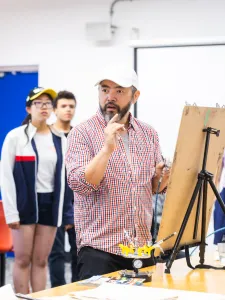 Students gather around an instructor giving an art demonstration in a studio space. Attention is focussed around an easel placed on top of a table. 