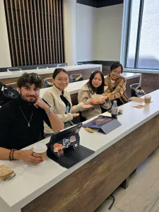 Four students sit at a table, laptops and food items in front of them. 