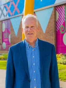 Photograph of David Binet wearing a blue blazer, standing in Butterfield Park on a sunny day.
