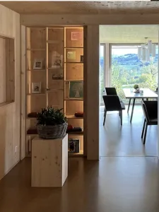 Photo of a room with wooden interiors. There are shelfs with decorative things on them. There is a table and a couple of chairs in the next room, which also has a wall to wall window.