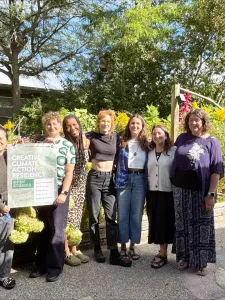 A group of seven people standing in front of a building with green bushes in the background