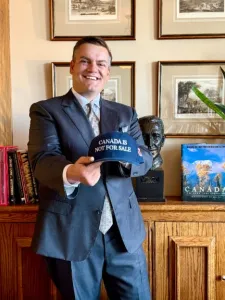Photo of a man wearing a blue suit and tie holding up a baseball cap that says Canada is not for sale, standing in front of a cabinet with bookshelf to the left, and photos hanging on the wall behind him.