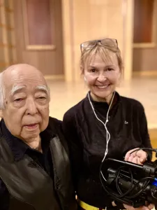 Japanese-Canadian man at left with a woman with blond hair and glasses on her head, holding a camera.