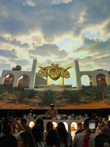 An event with a DJ performing on stage with a screen in the background displaying a desert scene, architectural structures and a golden bee in the center.