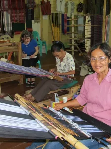 Three weavers working on a loom, woman in foreground smiling at camera wearing pink shirt