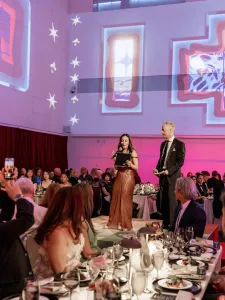 Gala Co-Chairs Amy Burstyn-Fritz and Jeff Hull stand on the centre stage addressing guests during the 2026 OCAD U Gala. Attendees seated at banquet tables surround the stage while large-scale projected visuals from Solarium by OCAD U alum Cell Driver illuminate the walls of the Great Hall.