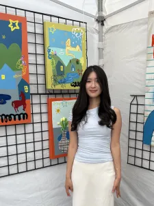 Woman with long black hair wearing pale blue blouse and white skirt standing among her paintings