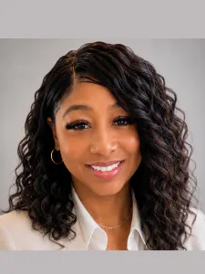 Woman with long dark curly hair, wearing white shirt and beige blazer