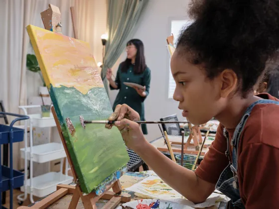 Young female teen is painting at an easel and an instructor is standing in the background.