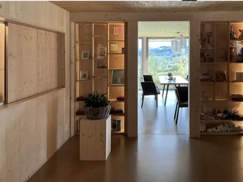 Photo of a room with wooden interiors. There are shelfs with decorative things on them. There is a table and a couple of chairs in the next room, which also has a wall to wall window.