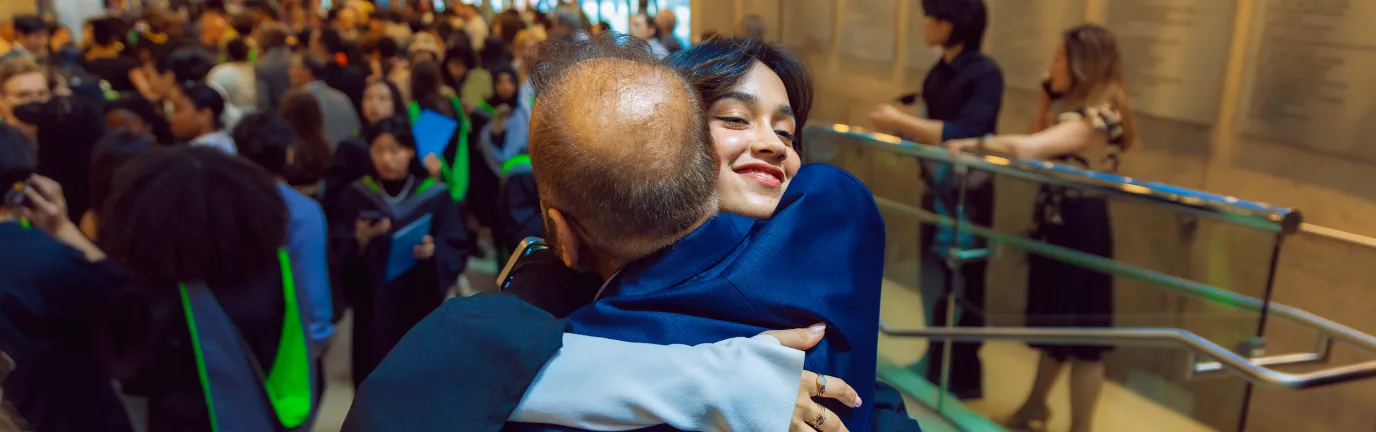 Two people in an embrace, with a group of people in graduation robes behind them.