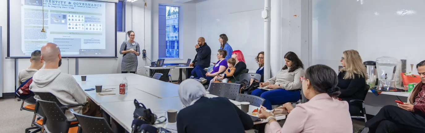 White room with a table in the centre, while people look towards a woman.
