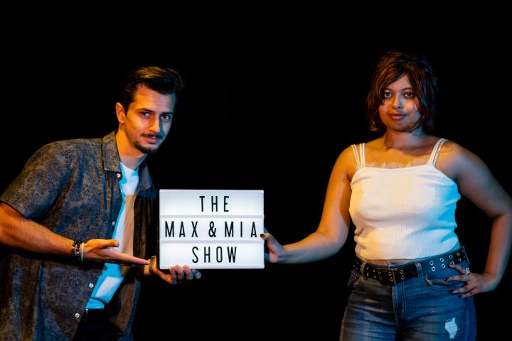 a young man and woman hold a small sign reading "The Max and Mia Show"