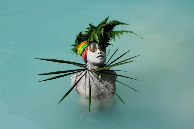 A photo of a person in a body of water with their skin painted white wearing head and neck pieces made of green fronds.. 