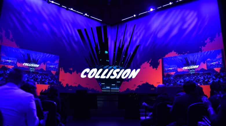 A blue toned photograph of a presentation hall with large screens with digital projections. The words Collision appears in the centre of the image.