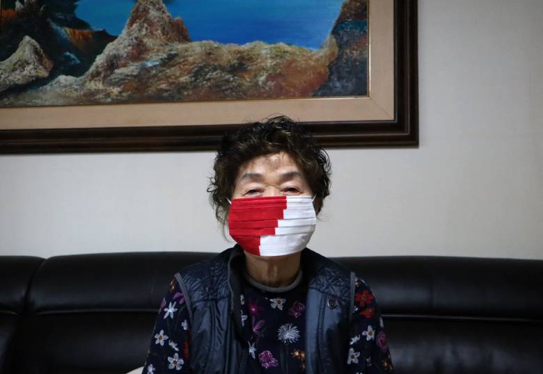 An Asisan woman wears a medical face mask sitting on a couch in a living room.
